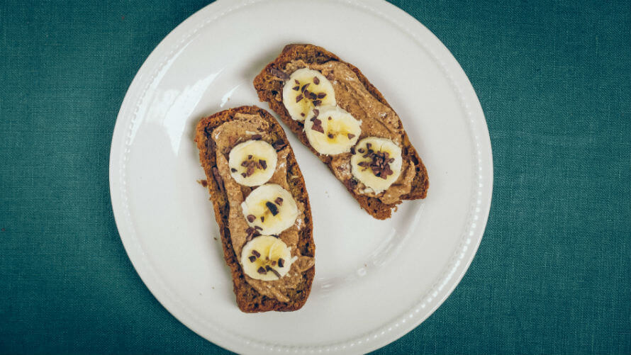 Two slices of sprouted banana bread on a plate topped with almond butter, sliced banana and cacao nibs