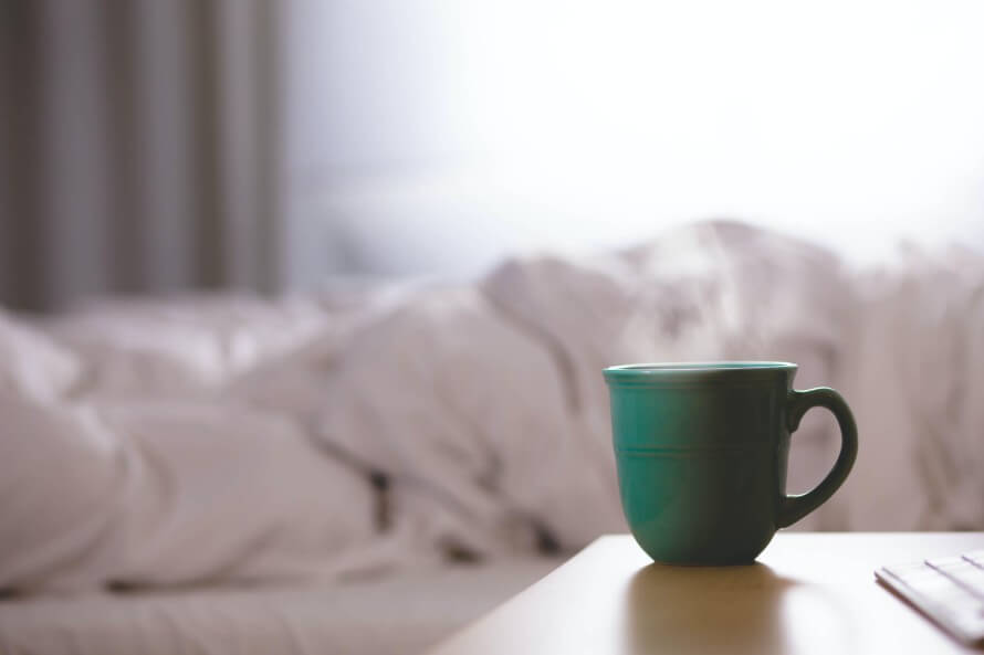 Bedroom. Steaming light green mug on a nightstand in foreground. White unmade bed before a window in background. 