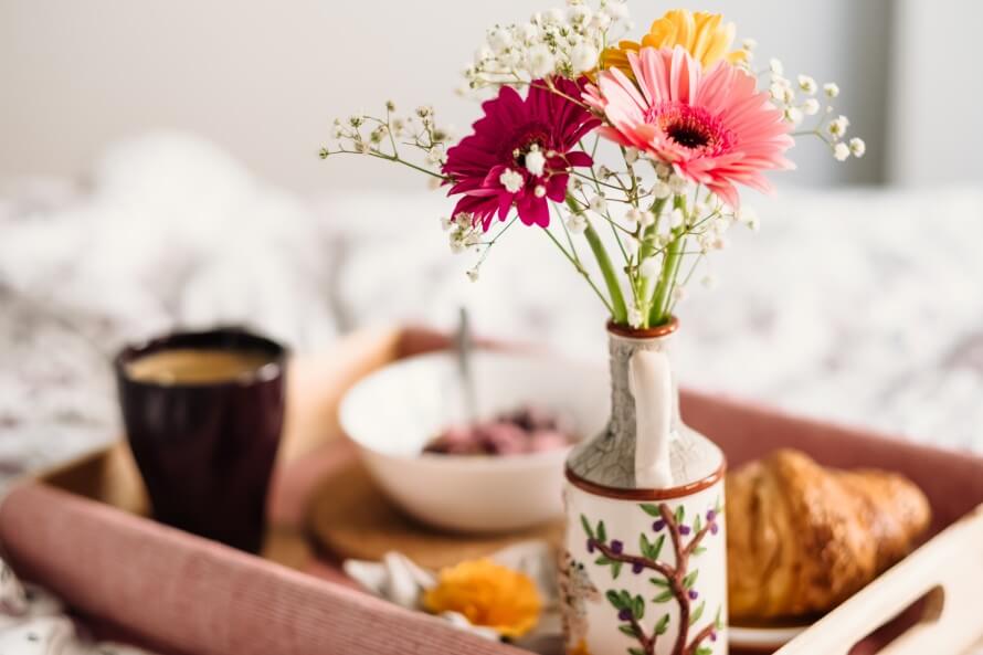 Breakfast tray on bed filled mug of coffee, bowl of granola, croissant, and a flower bouquet in a decorative ceramic. 