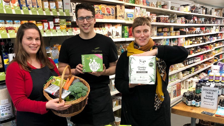 Hannah, Russell, and Rachel standing in shop in front of Organico BASICS shelves. Each holding various groceries from the range. 