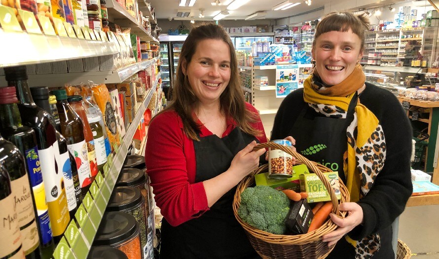 Hannah and Rachel standing beside shop shelves holding a basket filled with groceries from Organico BASICS range. 