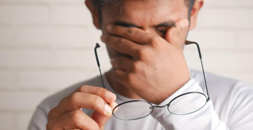 Tired man holding glasses in one hand. His other hand over his face and his fingers on either side of his eyes. 