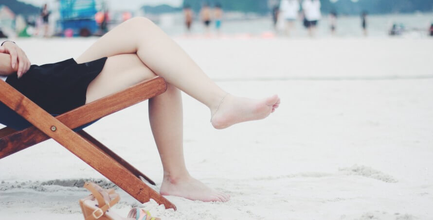 Lower body of fair-skinned woman in black dress sitting on a wooden beach chair on a sandy beach. Blurred bodies in the background.  