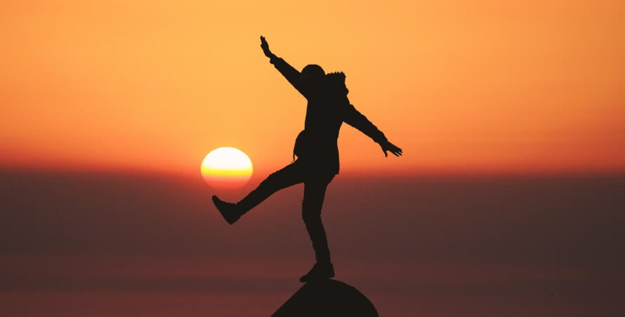 Silhouette of person in winter coat standing on point of rock. Kicking out one leg and one arm raised, the other extended back. Orange and red sunset in background.  