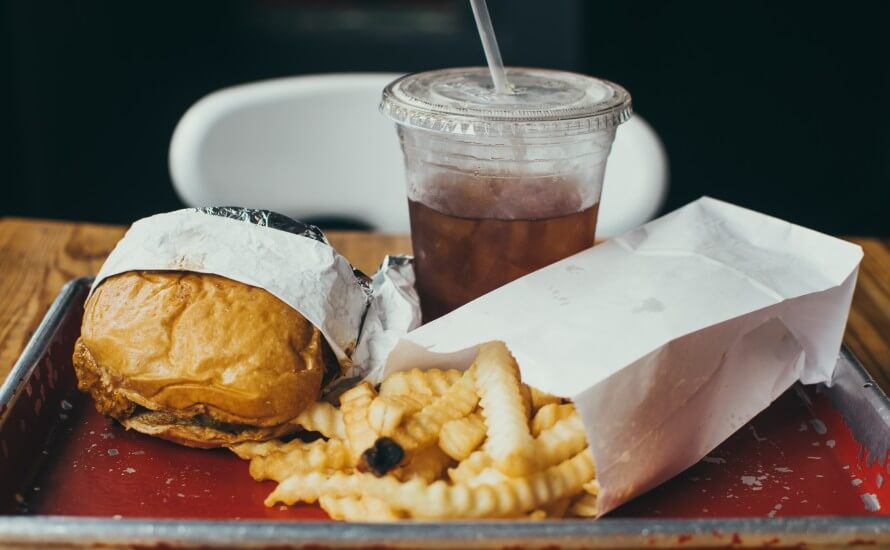 Fast food burger and curly cut fries un paper packaging and a soda in a plastic cup with lid and straw. All sitting on worn tray on a table in front of a white chair. 