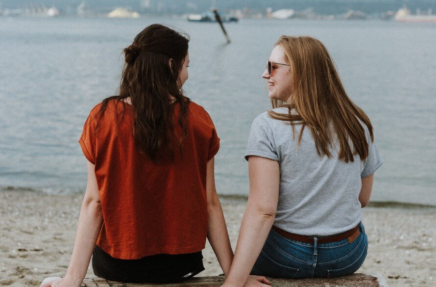 Two young women sitting on a log on the beach. Heads turned toward each other, smiling. 