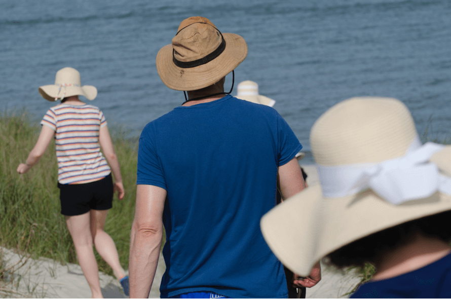 The backs of a group people all wearing sunhats and summer clothes. Walking on sunny day alongside a body of water. 
