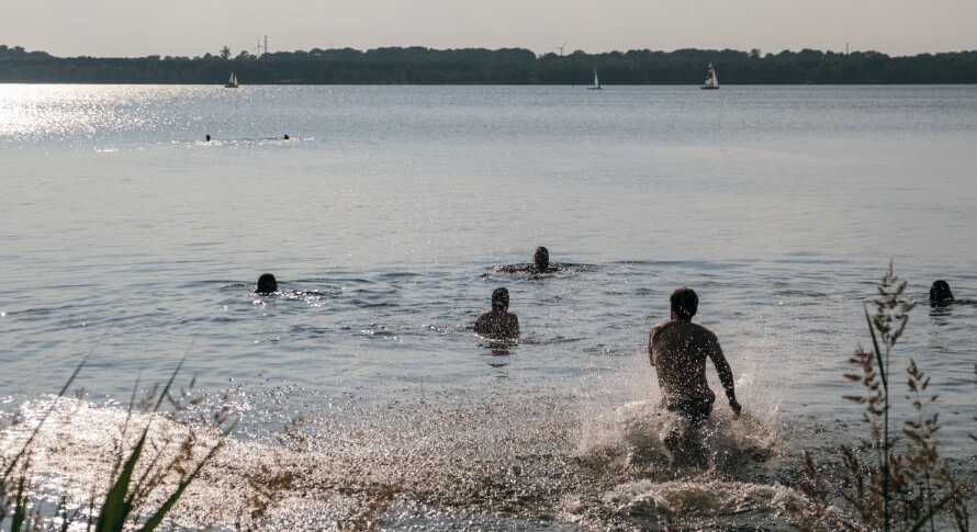 Group of people running into the ocean, splashing water. Sailboats in background. 