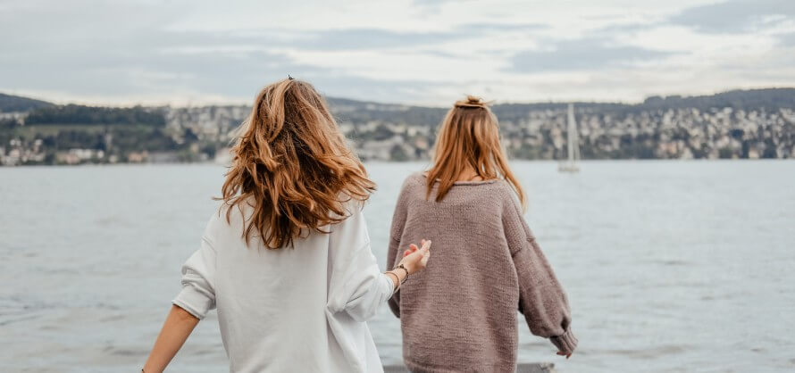 Two young women dressed in sweaters walking out onto a pier. Sailboat and houses in background. 