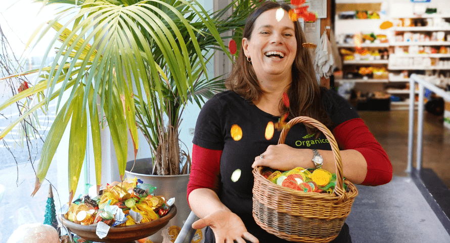 Hannah, smiling and wearing a black t-shirt adorned with the green Organico logo, stands in a well-lit grocery store beside a lush green potted plant. She's holding a woven basket filled with colorful, wrapped organic chocolate coins, suggesting a festive or seasonal celebration. The store shelves in the background are stocked with various products, highlighting the abundance of the organic market.