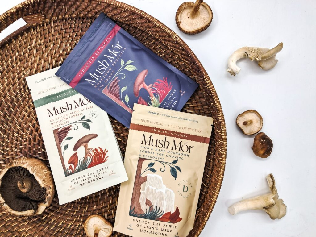 Three packets of Mush Mor adaptogenic mushrooms on a wicker platter. There are different types of fresh mushrooms scattered around.