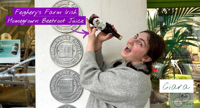 A smiling woman holds up a bottle of beetroot juice beside several award plaques on a wall, with overlaid text reading “Feighery’s Farm Irish Homegrown Beetroot Juice” and a handwritten label pointing to her that says “Ciara.” Green plants and a shop window appear in the background.